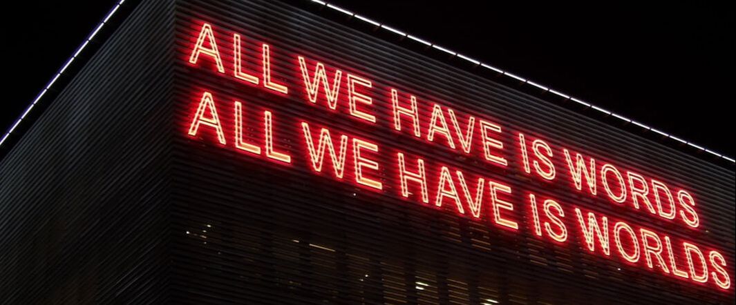 A neon sign on the side of a building reads ALL WE HAVE IS WORDS / ALL WE HAVE IS WORLDS